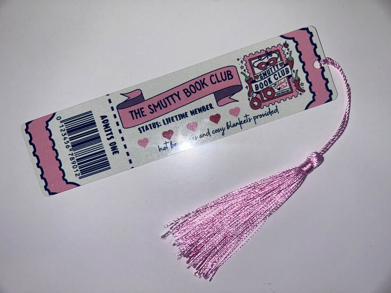 May include: A bookmark with a pink tassel featuring the text "THE SMUTTY BOOK CLUB". The bookmark has a ticket design with a barcode and the words "ADMITS ONE". The text "STATUS: LIFETIME MEMBER" is also visible.
