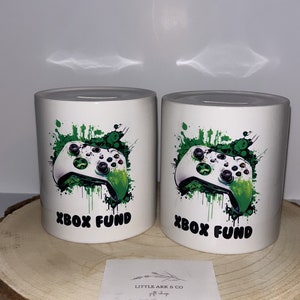 Puede incluir: Dos alcancías de cerámica blanca con gráficos de mando de Xbox verde y blanco y el texto "Xbox Fund". Las alcancías están sobre una superficie de madera.