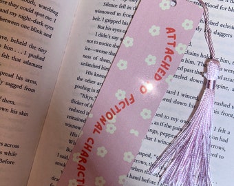 May include: A pink bookmark with the text "ATTACHED TO FICTIONAL CHARACTERS" and small white flower designs. It has a light purple tassel attached. The bookmark is placed in an open book.