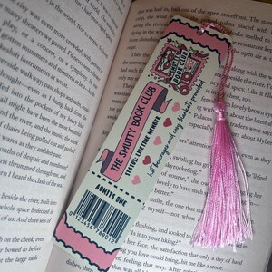 May include: A pink and white bookmark with the text "The Smutty Book Club" and "Admits One." It features a barcode and a pink tassel. The bookmark is resting on the pages of an open book.
