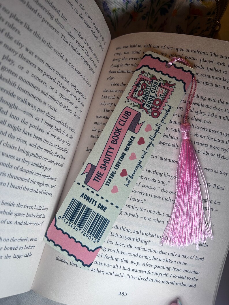 May include: A pink and white bookmark with the text "The Smutty Book Club" and "Admits One." It features a barcode and a pink tassel. The bookmark is resting on the pages of an open book.