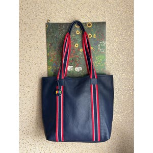 Op de afbeelding: Een donkerblauwe tote bag met rood-wit gestreepte banden. De tas heeft een klein, kleurrijk logo aan de voorkant.