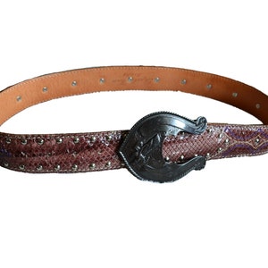 Può includere: Una cintura in pelle marrone con una fibbia argentata a forma di ferro di cavallo. La cintura è decorata con piccoli borchie argentate e un motivo a pelle di serpente.