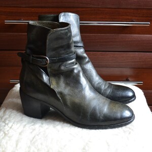 May include: A pair of dark green leather ankle boots with a chunky heel and a buckle strap. The boots are on a white surface.