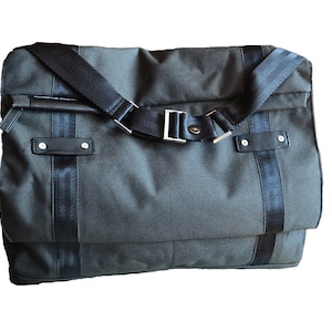 Puede incluir: Un bolso mensajero gris oscuro con correas y detalles en negro. El bolso tiene un cierre de solapa con hebilla y una correa de hombro ajustable. El bolso tiene una tela texturizada y detalles de cuero.