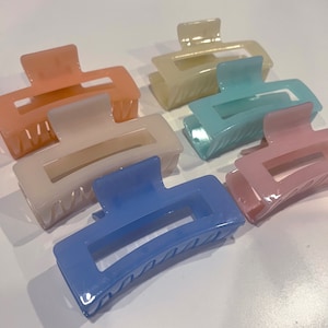 Puede incluir: Conjunto de seis pinzas para el pelo de plástico en colores pastel. Las pinces son de color rosa, azul y blanco. Cada pinza tiene una forma rectangular con la parte superior curvada y una pequeña abertura en el centro.