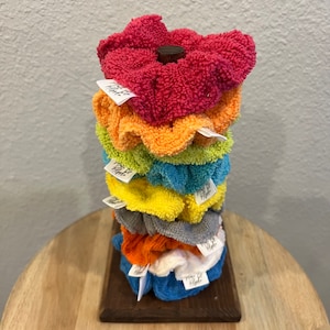 May include: A stack of colorful fabric scrunchies on a wooden stand. The scrunchies are in shades of red, orange, yellow, green, blue, and white. Each scrunchie has a small white tag attached to it.