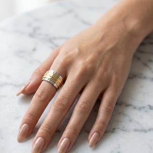 May include: A wide band ring featuring a central section of multiple thin, textured gold-toned wire coils flanked by two smooth, polished silver-toned metal bands on the top and bottom edges, creating a layered, multi-metal appearance.