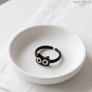 May include: A black open-band ring featuring a stylized cat face design with two pale yellow circular eyes. The ring is positioned inside a small white bowl. The text NOCTELLE JEWELRY is visible in the upper right corner of the image.