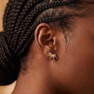 May include: A gold-toned ear stud featuring a sunburst or fan-shaped design with multiple thin, radiating rays. The earring is positioned on the earlobe, showcasing a metallic finish and a geometric, symmetrical arrangement of lines.