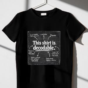 May include: A black t-shirt featuring a white graphic that resembles a chalkboard. The text reads: This shirt is. decodable. It includes linguistic notes like Closed Syllable, r-controlled, Digraphs, Latin based cas means down, Silent-e removed, and s says /z/.