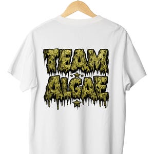 Peut inclure: Un t-shirt blanc à manches courtes avec un motif au dos portant le texte "TEAM ALGAE" dans une police vert olive texturée et dégoulinante. Le design comprend deux petites étoiles placées entre les mots et sous le texte.