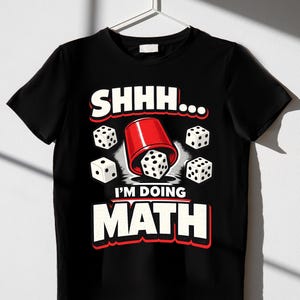 Peut inclure: Un t-shirt noir à manches courtes avec une impression graphique représentant un gobelet à dés rouge et des dés blancs. Le texte SHHH... I'M DOING MATH est écrit en lettres blanches grasses avec un contour rouge. Quatre dés blancs entourent le gobelet.