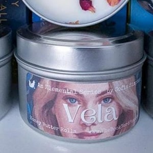 Peut inclure: Une boîte de bougie en argent avec une bougie blanche et des pétales de rose séchés. L'étiquette indique "An Elemental Series by Sofie Sims Vela Honey Butter Rolls www.thebooknerd.com"