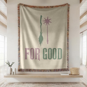 Peut inclure: Plaid beige avec des franges. Le plaid présente un balai vert et une baguette en forme d'étoile violette, avec les mots "FOR GOOD" en violet et vert.