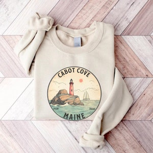 May include: A beige sweatshirt featuring a circular graphic of a lighthouse on a rocky island with a sailboat in the water. The graphic reads "Cabot Cove Maine" and states the population as 3650.