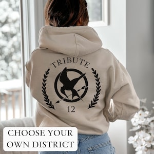 May include: A beige hoodie with a black graphic on the back. The graphic features a mockingjay bird, a circle, and arrows, with the text "TRIBUTE" above and "12" below. The text "CHOOSE YOUR OWN DISTRICT" is at the bottom of the image.