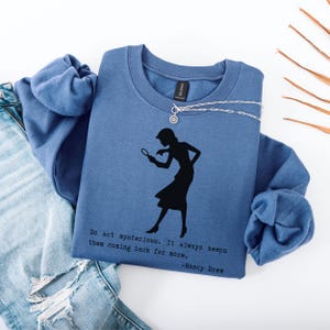 May include: A blue sweatshirt with a silhouette of a woman holding a magnifying glass. The text "Do act mysterious. It always keeps them coming back for more. -Nancy Drew" is printed on the sweatshirt.