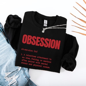 May include: A black long-sleeve sweatshirt featuring the red text OBSESSION, the phonetic spelling ob-ses-sion (in), and a dictionary-style definition: 1. A relentless attachment to an idea, feeling, or emotion: often paired with mental strain and physical unease.