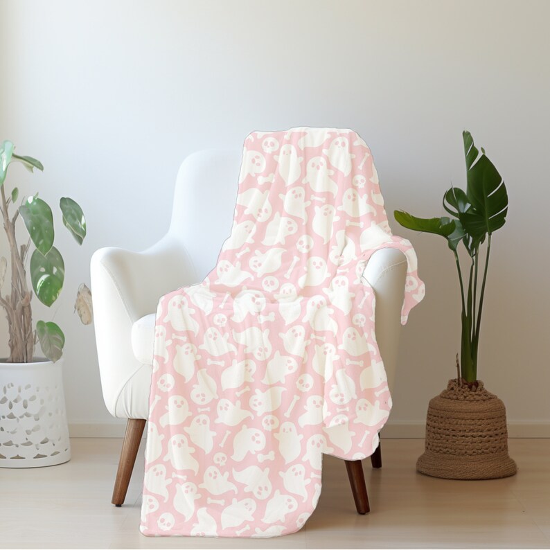 Pu&ograve; includere: Una coperta in pile rosa con un motivo di fantasmi e ossa bianche. La coperta &egrave; drappeggiata su una poltrona bianca.