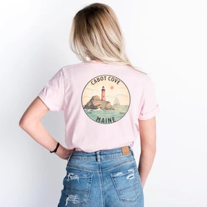 Puede incluir: Una camiseta rosa con un gr&aacute;fico en la parte posterior de un faro en una isla rocosa con el texto "CABOT COVE" y "MAINE".