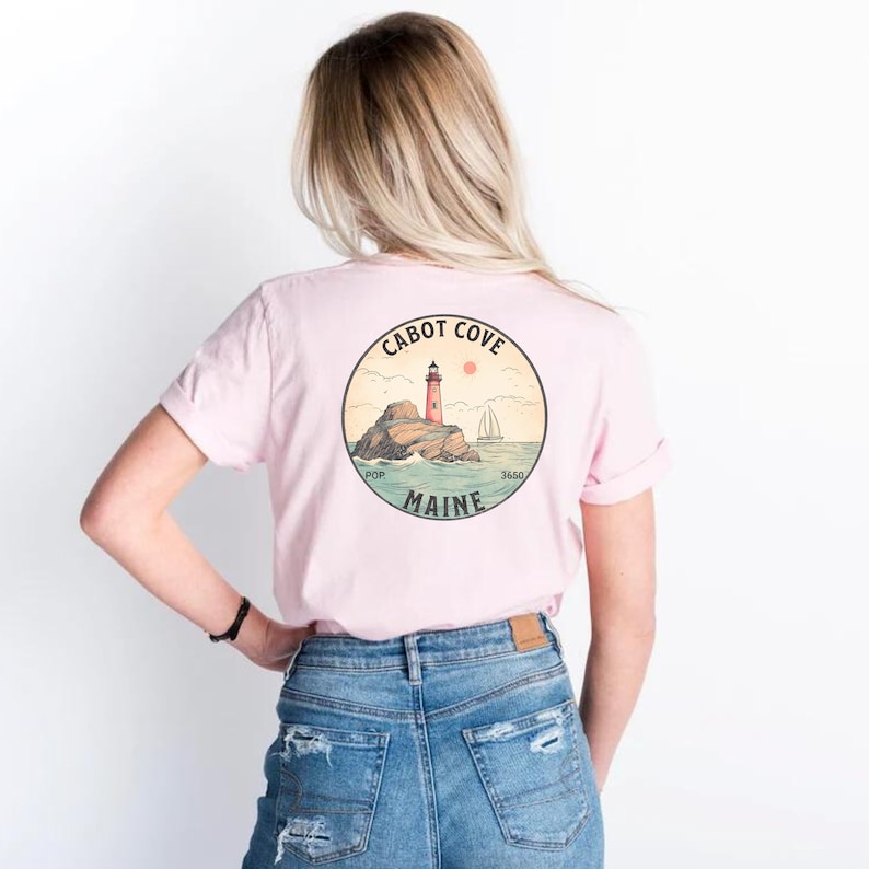 Puede incluir: Una camiseta rosa con un gr&aacute;fico en la parte posterior de un faro en una isla rocosa con el texto "CABOT COVE" y "MAINE".