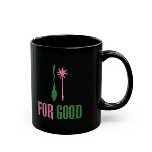 May include: Black ceramic mug featuring a pink and green design with a witch's broom, a star-shaped wand, and the words "FOR GOOD" in pink and green. The mug has a curved handle and a glossy finish.