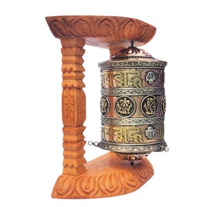 May include: A wooden prayer wheel with intricate carvings and a copper-colored metal cylinder. The prayer wheel is mounted on a wooden stand with detailed carvings. The cylinder has engraved text and decorative elements.