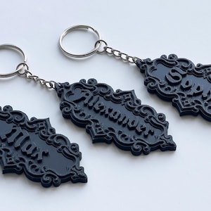 May include: Three black keychains with ornate, decorative frames and silver key rings. Each keychain has a name engraved in a cursive font. The names are Max, Alexander, and Sofia.