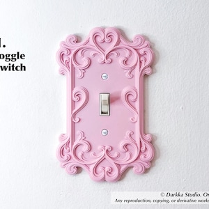 Puede incluir: Una cubierta de interruptor basculante rosa con un dise&ntilde;o ornamental de volutas. La cubierta tiene forma rectangular con un interruptor central y est&aacute; montada en una pared blanca. El texto "1. toggle switch" est&aacute; en la esquina superior izquierda.