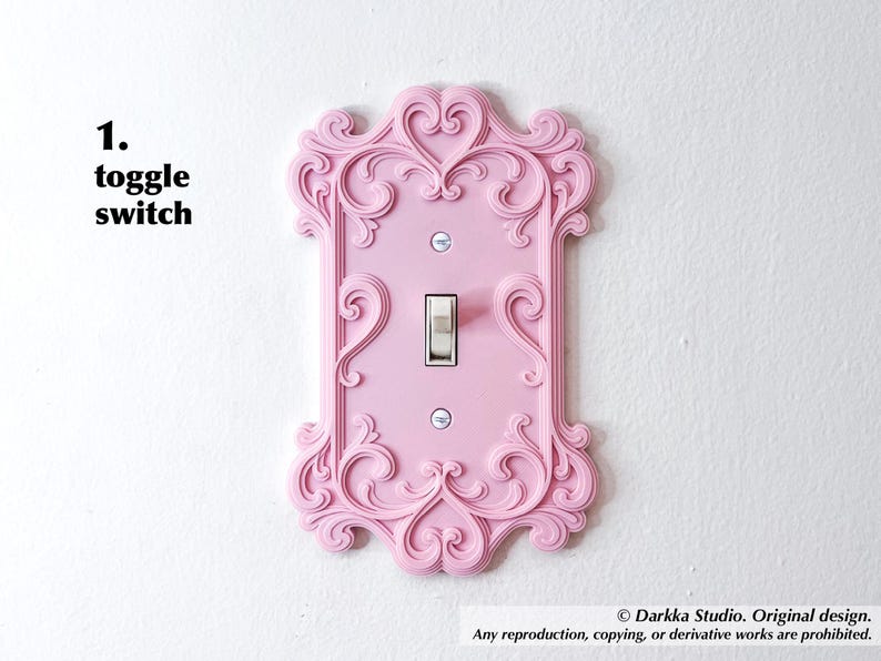 Puede incluir: Una cubierta de interruptor basculante rosa con un dise&ntilde;o ornamental de volutas. La cubierta tiene forma rectangular con un interruptor central y est&aacute; montada en una pared blanca. El texto "1. toggle switch" est&aacute; en la esquina superior izquierda.