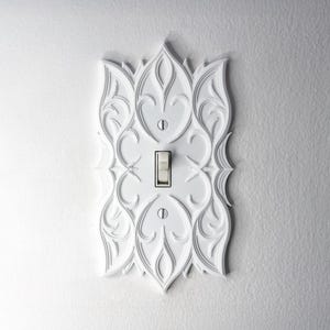 Puede incluir: Cubierta de interruptor de luz decorativa blanca con un diseño floral ornamentado. La cubierta tiene un interruptor basculante en el centro y está montada en una pared blanca. El diseño incluye patrones detallados y arremolinados.