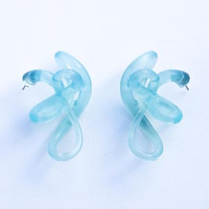 May include: A pair of light blue resin earrings with a knotted design. The earrings are made of a translucent material and have a simple, modern look.