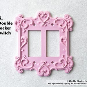 Puede incluir: Una placa de interruptor basculante doble rosa con detalles ornamentados y arremolinados y acentos en forma de coraz&oacute;n. La placa tiene dos aberturas para interruptores y el texto "4. Double rocker switch".