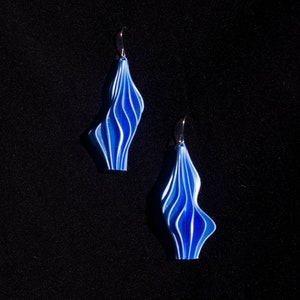 Może przedstawiać: Para niebieskich i białych kolczyków z falistym, złożonym wzorem. Kolczyki są wykonane z lekkiego materiału i mają srebrny haczyk.
