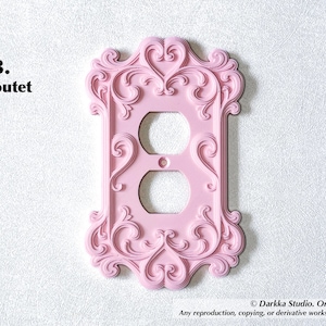 Puede incluir: Una cubierta de enchufe rosa y ornamentada con un dise&ntilde;o decorativo y ondulado. La cubierta tiene dos agujeros para los enchufes y est&aacute; montada en una pared blanca texturizada. El texto "3. outet" est&aacute; en la esquina superior izquierda.