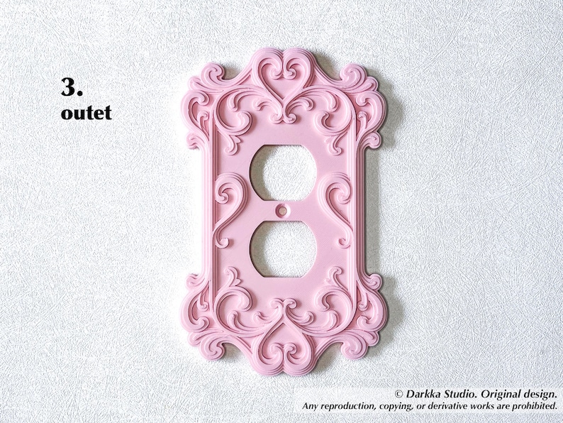 Puede incluir: Una cubierta de enchufe rosa y ornamentada con un dise&ntilde;o decorativo y ondulado. La cubierta tiene dos agujeros para los enchufes y est&aacute; montada en una pared blanca texturizada. El texto "3. outet" est&aacute; en la esquina superior izquierda.