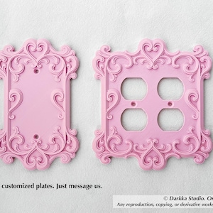 Puede incluir: Dos placas decorativas rosas para interruptores de luz con adornos de volutas y detalles en forma de coraz&oacute;n. Una es una placa de un solo interruptor y la otra tiene cuatro orificios para enchufes. El texto dice: "Ofrecemos placas personalizadas. Cont&aacute;ctenos."