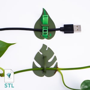 May include: Green leaf-shaped cable organizers with a USB cable and a plant stem. The organizers are designed to hold cables and plant stems, with a green clip to secure them. The image also includes the text "STL".