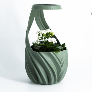 K&ouml;nnte beinhalten: Ein salbeigr&uuml;ner, 3D-gedruckter Pflanztopf mit einem einzigartigen, wirbelnden Design. Der Pflanztopf enth&auml;lt eine kleine wei&szlig; bl&uuml;hende Pflanze mit gr&uuml;nen Bl&auml;ttern. Der Pflanztopf hat einen Griff und den Text "Factorian Designs".