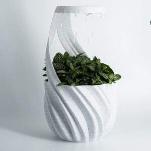 K&ouml;nnte beinhalten: Ein moderner, grauer Pflanztopf mit einem einzigartigen, geschwungenen Design, gef&uuml;llt mit &uuml;ppigen gr&uuml;nen Pflanzen. Der Pflanztopf hat ein strukturiertes, wirbelndes Muster und eine griffartige Struktur. Design von Factorian Designs.