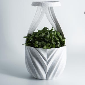 K&ouml;nnte beinhalten: Ein grauer, strukturierter H&auml;ngepflanzentopf mit geometrischem Design, gef&uuml;llt mit gr&uuml;nen Pflanzen. Der Pflanzentopf hat eine kreisf&ouml;rmige Oberseite und einen tropfenf&ouml;rmigen K&ouml;rper. Design von Factorian Designs.