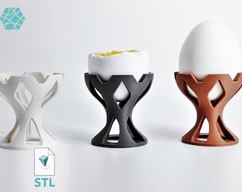 May include: Three egg cups in white, black, and brown, each featuring a geometric, open-lattice design. The black cup holds a cracked egg, while the brown cup holds a whole white egg. The white cup is empty. Each cup has a wide, circular base.