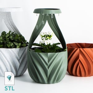 May include: Three decorative planters in shades of gray, green, and orange. The gray planter has a handle and is filled with green plants. The green planter has a unique design and holds white flowers. The orange planter has a textured surface.