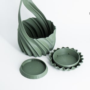 K&ouml;nnte beinhalten: Ein salbeigr&uuml;ner, 3D-gedruckter Pflanztopf mit einem einzigartigen, wirbelnden Design und einem Griff. Der Pflanztopf wird von zwei passenden gr&uuml;nen, kreisf&ouml;rmigen Teilen begleitet, wahrscheinlich einem Boden und einem Deckel. Das Logo von Factorian Designs befindet sich oben rechts.