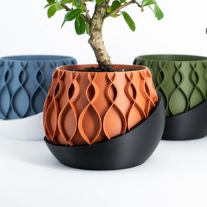 May include: Three decorative plant pots in blue, orange, and green, each with a unique, wave-like pattern. The pots sit on black, curved bases. A small bonsai tree is planted in the orange pot. The Factorian Designs logo is in the upper right corner.
