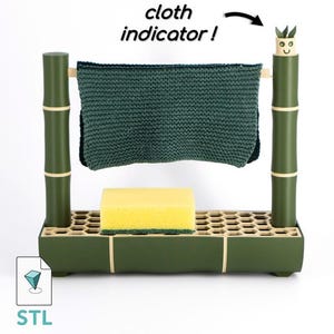 Könnte beinhalten: Ein grüner Geschirrtrockner im Bambus-Stil mit einem dunkelgrünen Tuch und einem gelben Schwamm. Der Ständer hat eine wabenförmige Basis und den Text "cloth indicator!". Das Design beinhaltet dekorative Elemente.