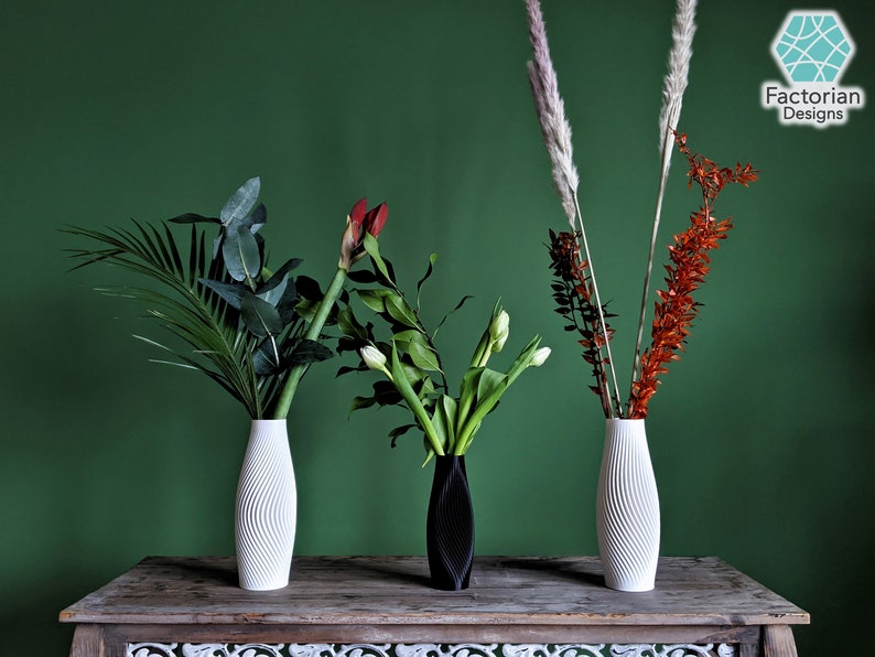 Puede incluir: Tres jarrones de cer&aacute;mica blanca con diferentes patrones texturizados, que contienen varias flores y plantas verdes. Los jarrones est&aacute;n dispuestos sobre una mesa de madera con una superficie estampada. El fondo es una pared verde s&oacute;lida.