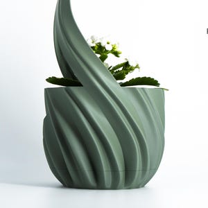 K&ouml;nnte beinhalten: Ein salbeigr&uuml;ner, 3D-gedruckter Pflanzentopf mit einem einzigartigen, wirbelnden Design. Der Topf enth&auml;lt eine kleine Pflanze mit wei&szlig;en Bl&uuml;ten und gr&uuml;nen Bl&auml;ttern. Das Logo von Factorian Designs ist sichtbar.