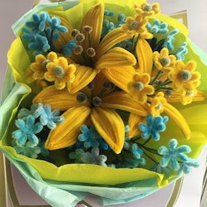以下が含まれることがあります： 手作りの花のブーケ。大きな黄色のユリと、より小さな黄色と青の花が特徴です。アレンジメントは、黄色、青、ピンクの薄紙の層で包まれており、明るく陽気なディスプレイを作成しています。